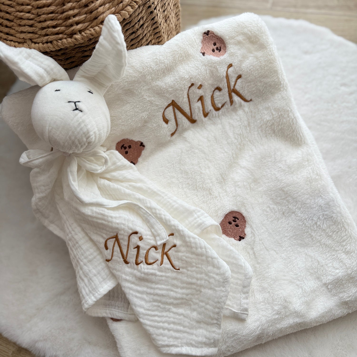 White baby blanket with embroidered name 'Nick' and a soft toy on a wooden surface.