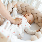 Baby Natur beige braided crib bumper made of soft, natural materials, protecting a baby in a white cherry-print sleep sack.