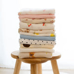Stack of Baby Natur 100% cotton muslin cloths with various cute prints like cherries, bears, dogs, and polka dots.