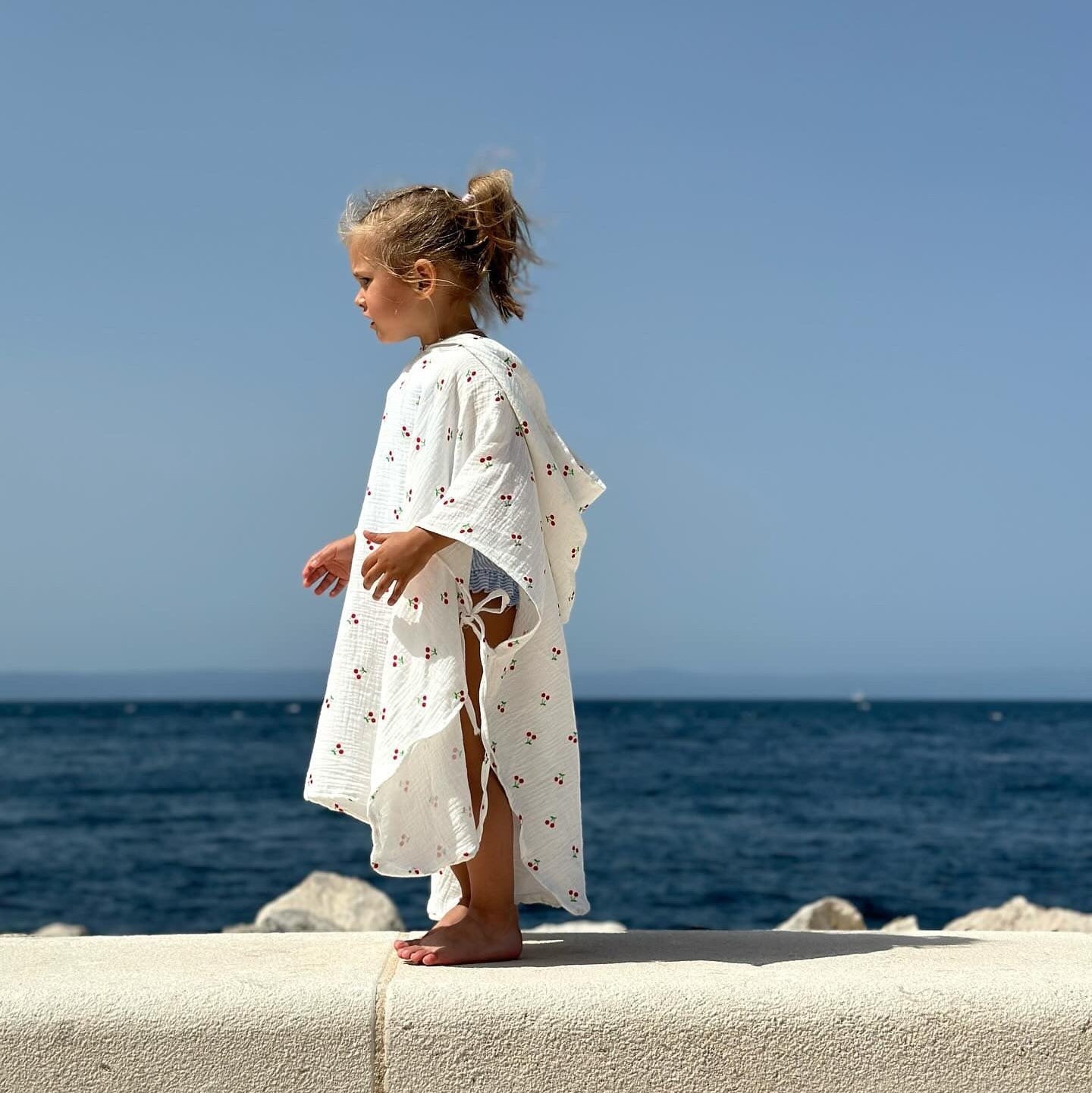Baby Natur white muslin poncho with cherry print, featuring side ties and a hood. Perfect for beach or pool.