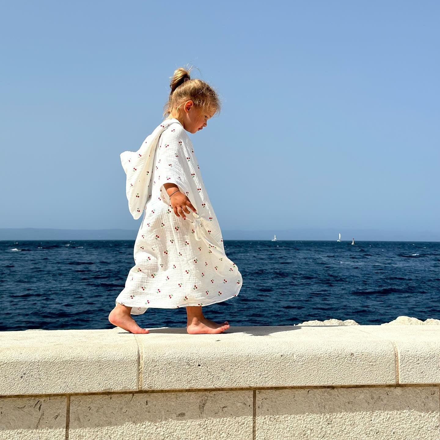Baby Natur white muslin poncho with red cherry print, worn by a young girl walking by the sea.